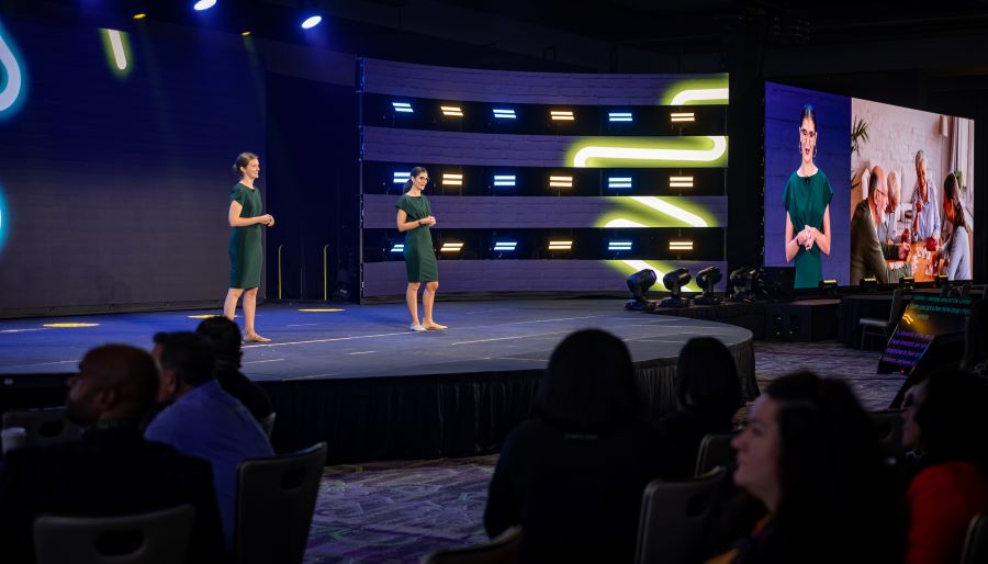 Event stage with a speaker, dynamic lighting, and a large presentation screen, with an audience in the foreground.