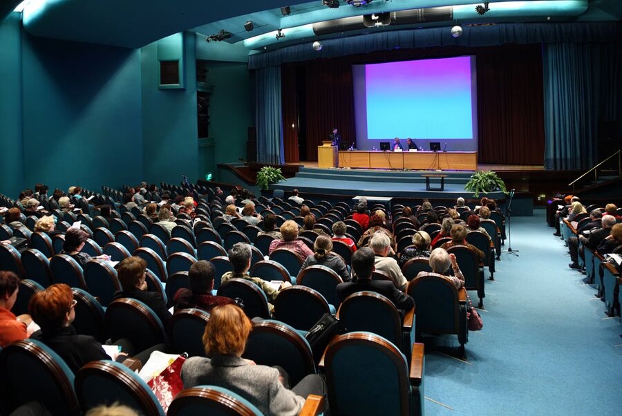 An auditorium filled with people in the seats, an event on stage with a large screen on display.