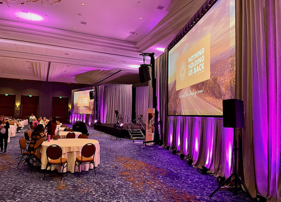 Ballroom venue with purple uplights, large presentation screens, and speakers.