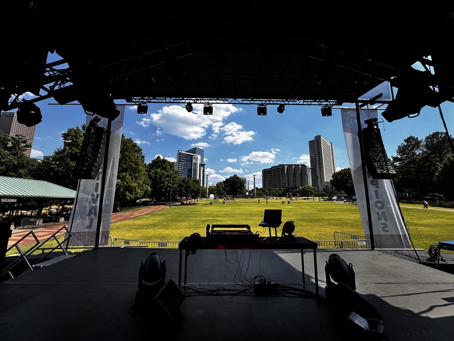 View from a stage, showing a setup with speakers, lighting, and a console overlooking an open outdoor park with a city skyline and a bright blue sky in the background