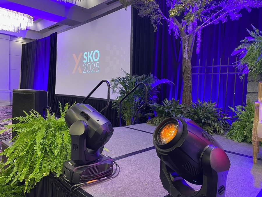 Ballroom event stage setup featuring vibrant purple lighting, a projection screen with 'XSKO 2025' branding, greenery, and professional stage lighting equipment