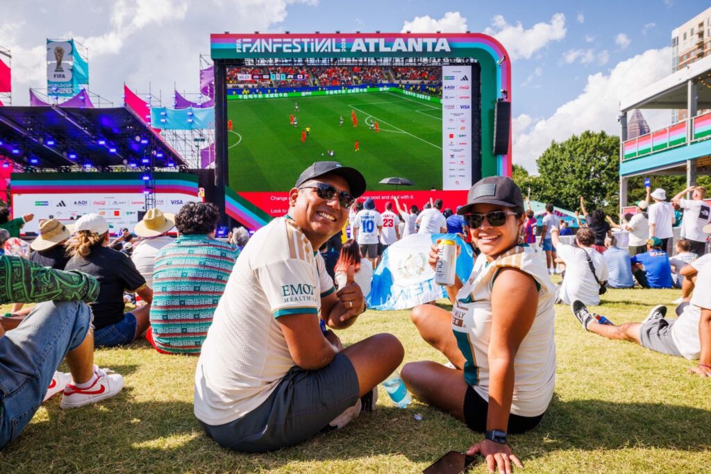 Man and woman smiling while sitting in the grass watching soccer game at the FIFA Fan Festival